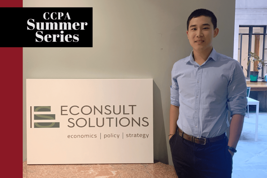 Ken Lin smiles next to an Econsult sign that says Econsult Solutions: Economics, Policy, Strategy.