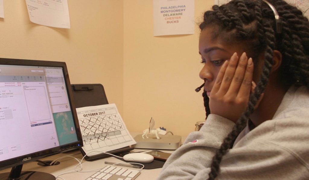 a young woman working a helpline sits at a desk, chin propped in one hand, looking dejected