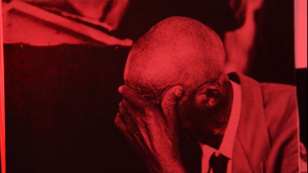 A close-up of a person seated and leaning forward, covering their face with one hand; the scene is bathed in an intense red light.