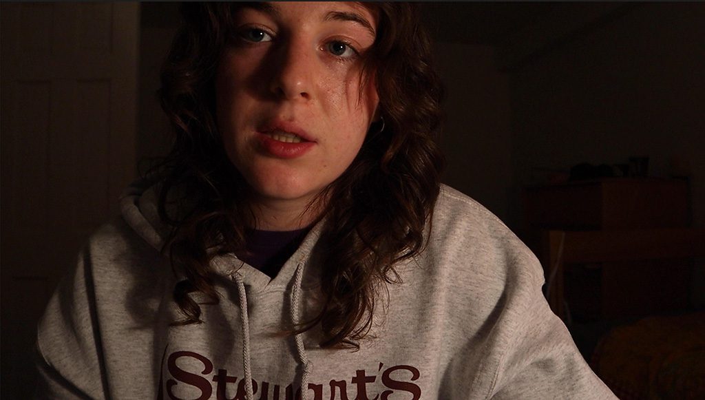 A close-up of a young person with wavy brown hair and a neutral expression, seated indoors against a dimly lit background; they wear a light-gray hoodie with partially visible maroon text and look slightly toward the camera.