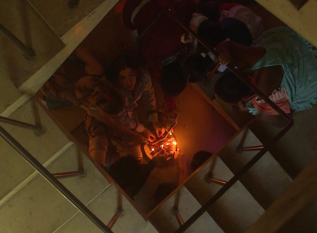 Overhead view down a square stairwell showing several people gathered on a landing around a small lit candle or diya; the warm light illuminates their faces and hands while the surrounding stairs remain dim.