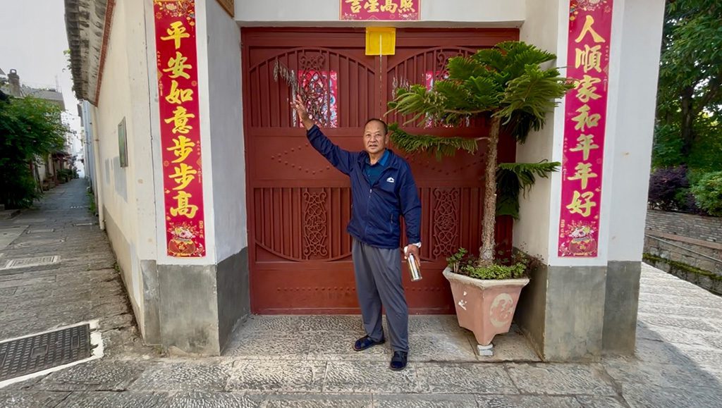  Zhang Canbao standing in front of a closed wooden gate decorated with red and gold vertical banners. A potted plant sits to their right and a narrow alley runs along the left side of the building.