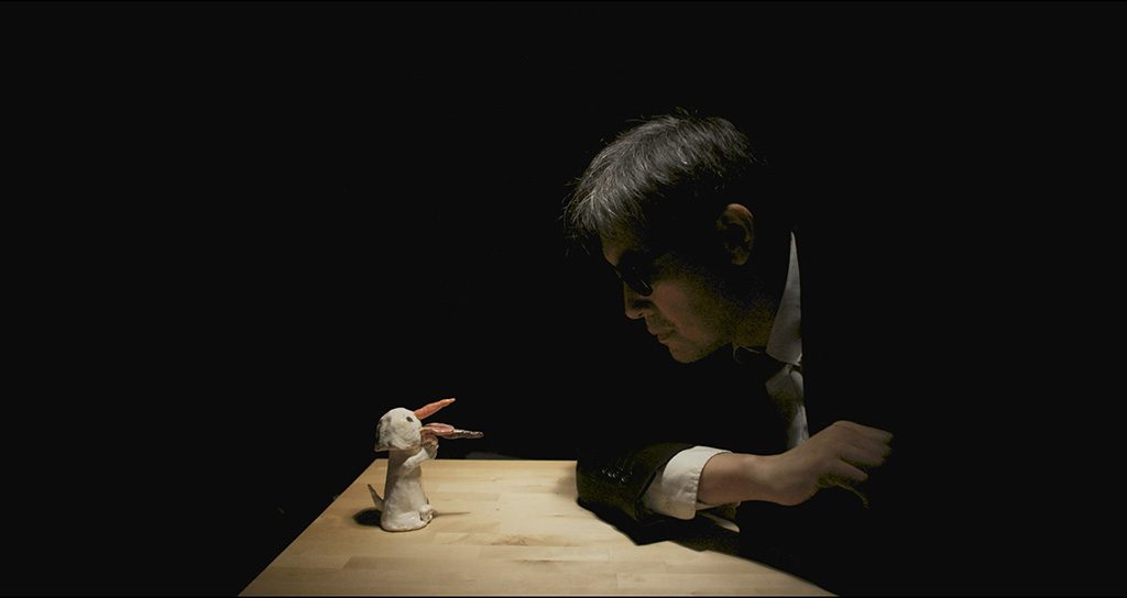 A dimly lit scene showing a person in a suit leaning over a table and staring at a small clay or puppet-like bird with its beak open; the background is black and a single light highlights the table and figures.