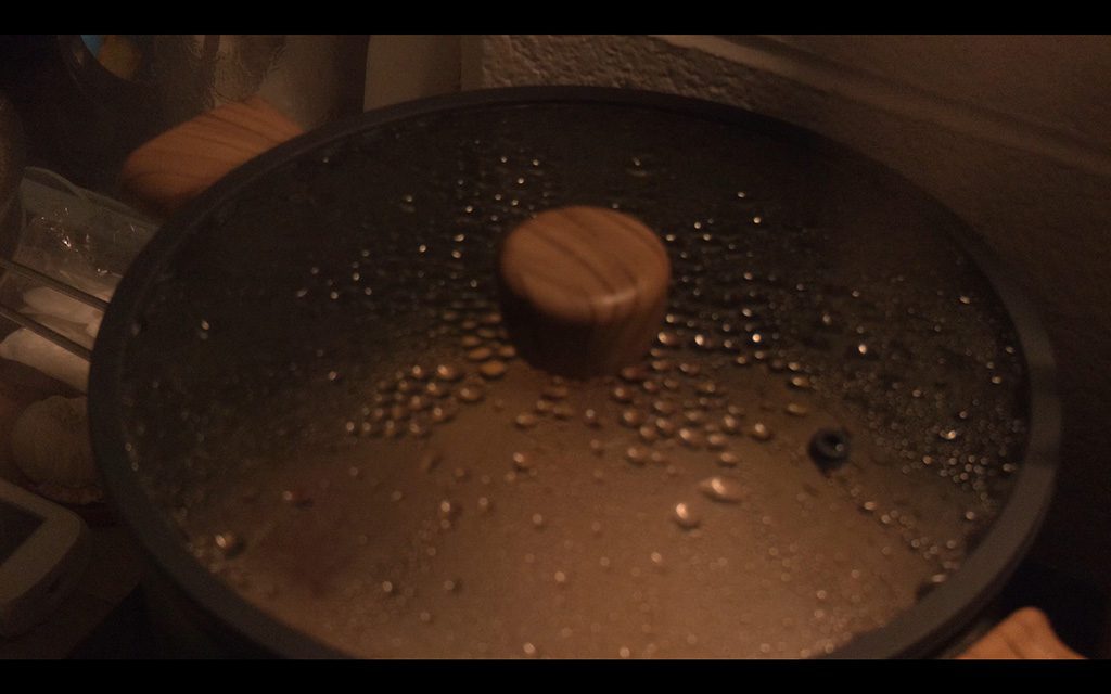 Close-up of a pot lid covered in water droplets with a wooden knob at the center; dim, warm lighting casts soft shadows across the metal surface.