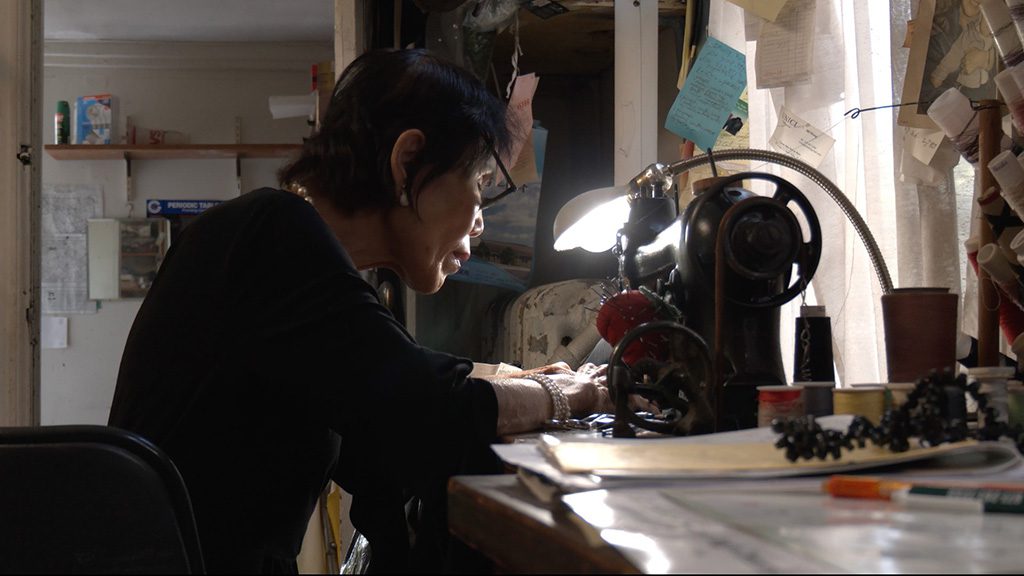 A person seated at a workbench, leaning forward and focused on a vintage sewing machine under a bright task lamp; papers and small tools are scattered on the table, and shelves and supplies fill the dimly lit workspace.