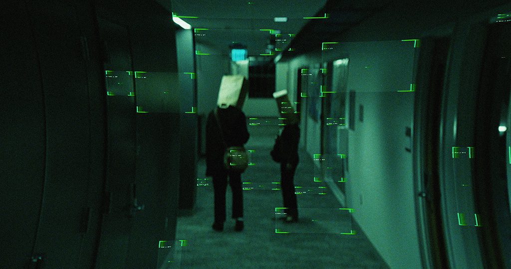 Two people standing in a dim, green-tinted hallway viewed through a digital targeting overlay; both wear paper bags over their heads and carry small bags, one facing slightly toward the other. The corridor has doors and recessed ceiling lights, and the image has a grainy, low-light look.