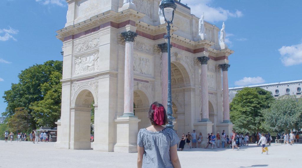 Person with short red hair and a striped shirt stands with their back to the camera, facing a large ornate stone arch in a sunny plaza; people stroll nearby under a blue sky.